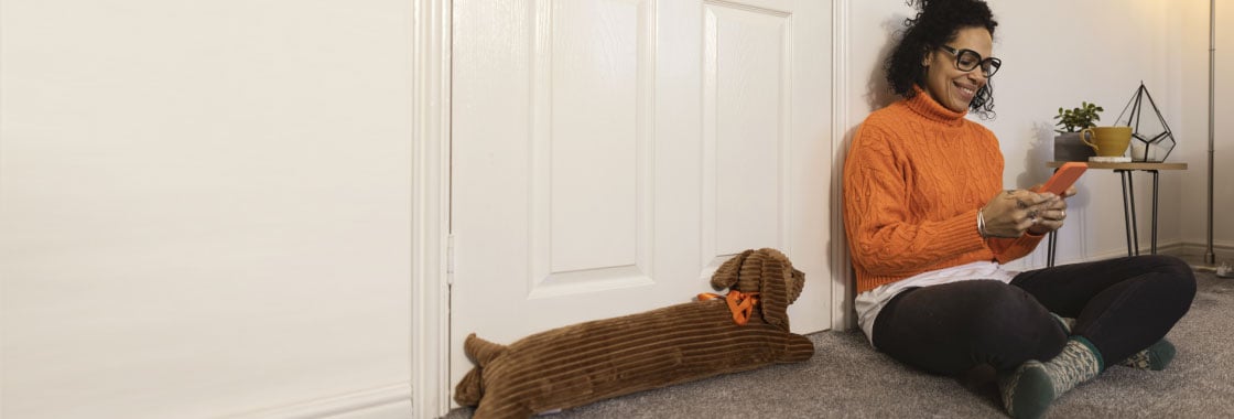 Smiling woman looking at her phone sits on the floor next to a door that has a woolly door stop in the shape of a dog.