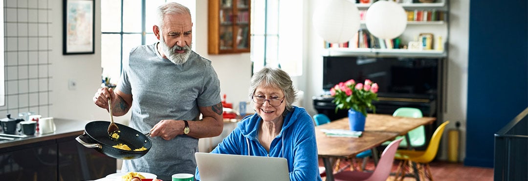 A man holds a pan of eggs while looking at his wife’s laptop