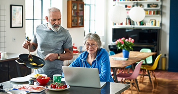 A man holds a pan of eggs while looking at his wife’s laptop